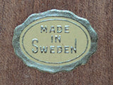 A vintage oval metal seal with a scalloped edge, featuring the text 'MADE IN SWEDEN' embossed in the center, resting on a brown wooden surface.